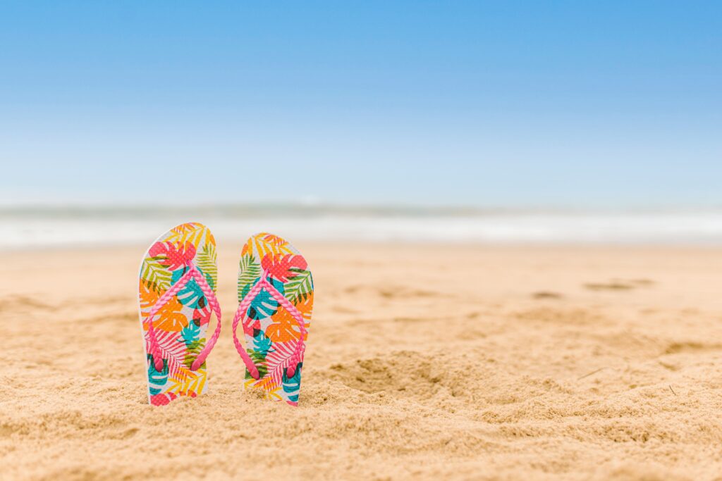 Vibrant patterned flip flops standing on a sandy beach with a clear blue sky.